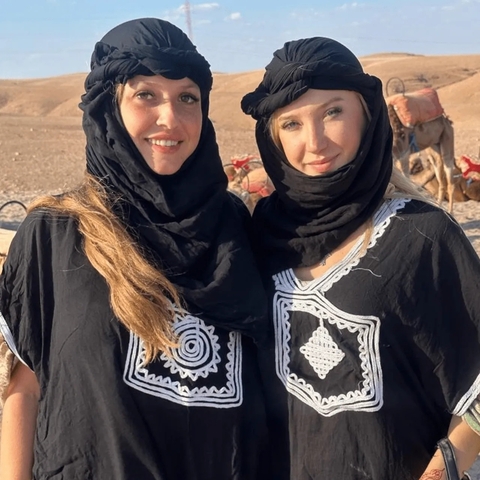      Two women in traditional Moroccan attire posing in a desert setting.
  