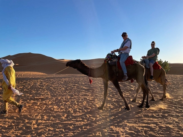 People riding camels in the desert with dunes in the background.