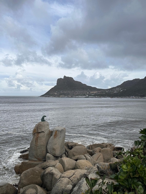       A coastal landscape with a statue on a rock and mountains in the background.
  