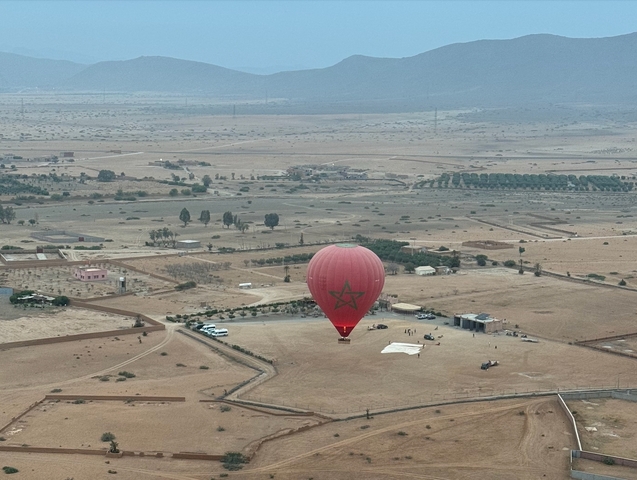       Red hot air balloon landing in a desert landscape with fields and mountains.
  