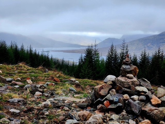 A cairn on a hill overlooking a loch with forested surroundings.