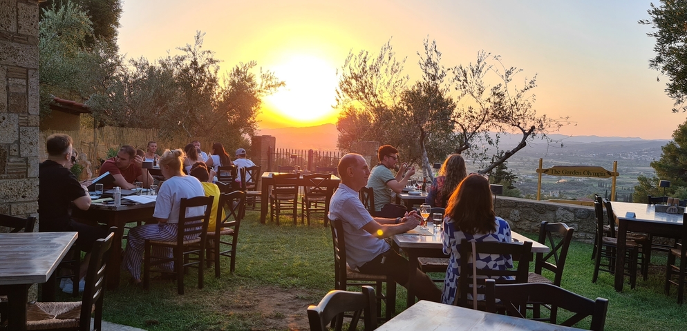       Open-air restaurant with people dining and watching the sunset over a hilly landscape.
  