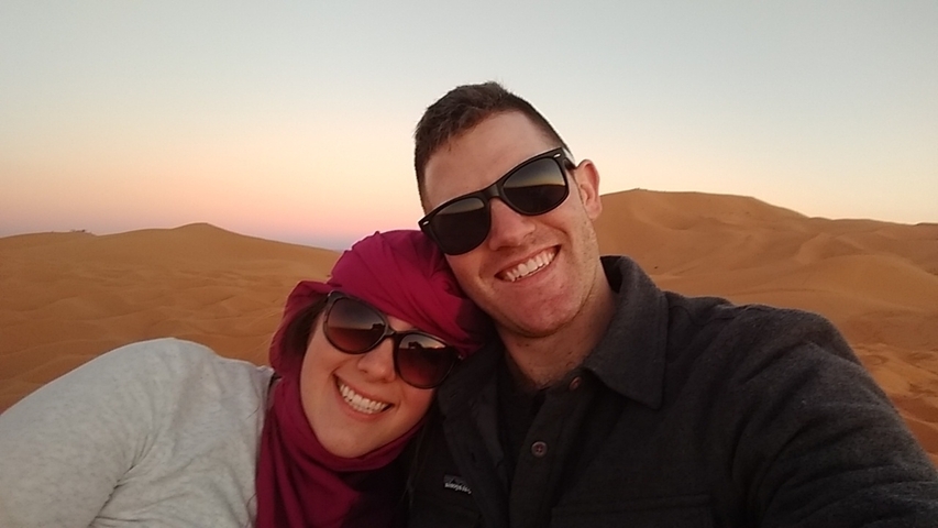 Couple smiling in a desert landscape with sunset in the background.