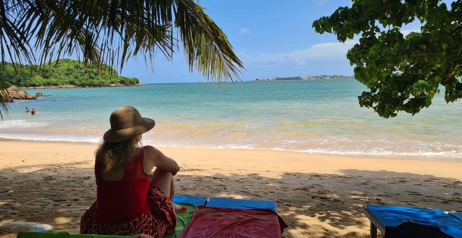 Woman sitting on a colorful mat on a sandy beach under palm trees.