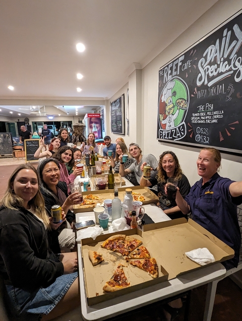       Group of people enjoying a meal with pizza boxes and drinks inside a casual restaurant.
  