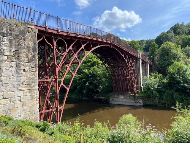 A historic red iron bridge over a river surrounded by greenery.