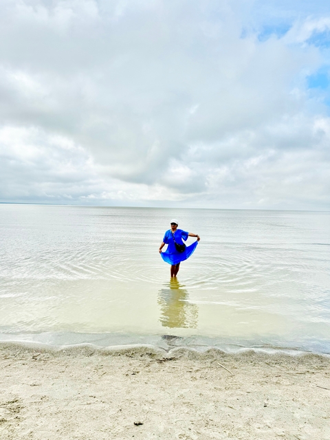       A person in blue standing in calm water with a cloudy sky.
  