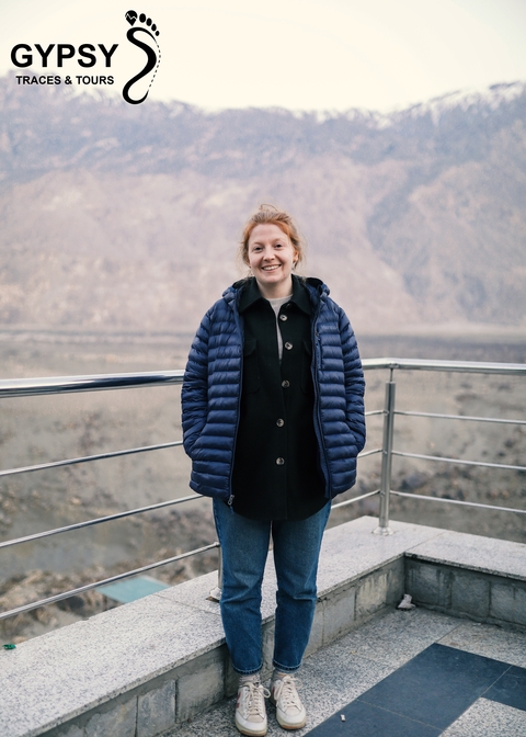       Person wearing a jacket standing on a balcony with mountains behind.
  