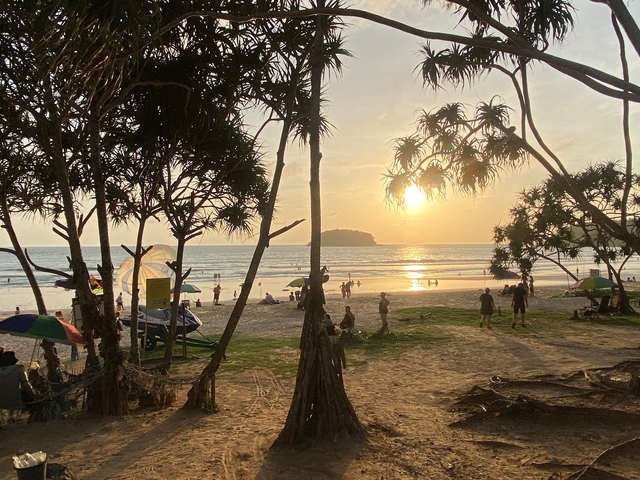       Sunset on a beach with people relaxing and trees in the foreground.
  