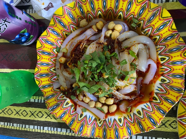 Bowl of spicy noodles with garnish, placed on a colorful tablecloth.
