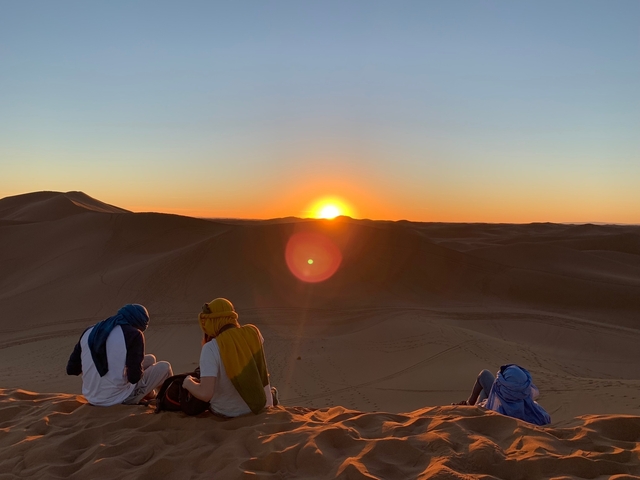       Two people sitting on sand dunes watching a sunset.
  