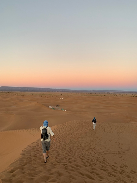 Two people walking on desert dunes during sunset.