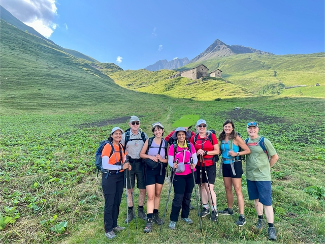       A group of hikers posing in a green valley with mountains in the background, under a blue sky.
  