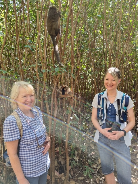       Two women smiling with a lemur in a forest setting.
  