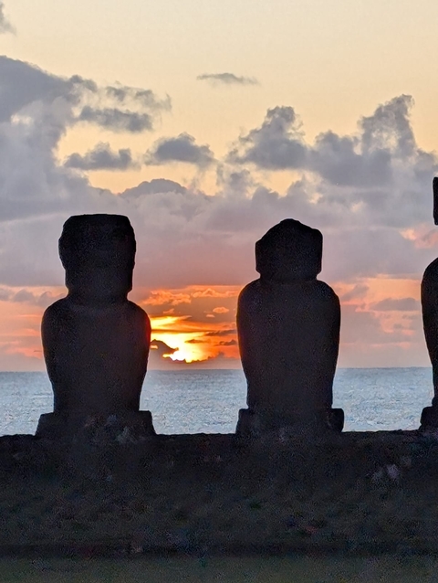       Moai statues overlooking a sunset by the ocean.
  