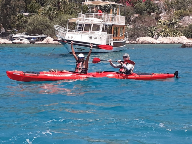 Two people kayaking in a bright blue sea with a boat in the background.