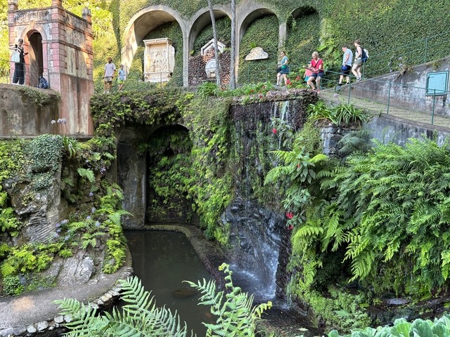       People walking across a mossy bridge in a lush garden.
  