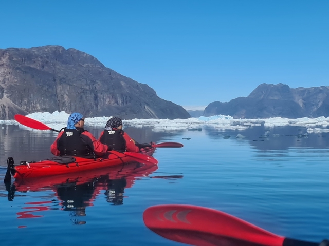      Two people kayaking among icebergs in a calm sea.
  
