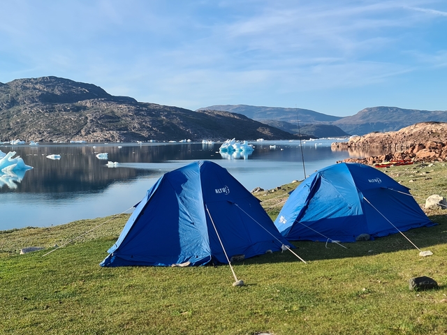       Two tents set up on grass with icebergs and mountains in the background.
  