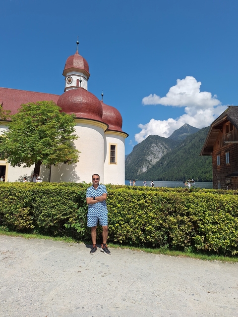       Man standing by a hedge with a picturesque church and mountain view.
  