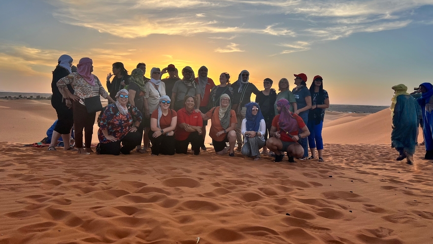       A large group of people posing together in the desert at sunset.
  