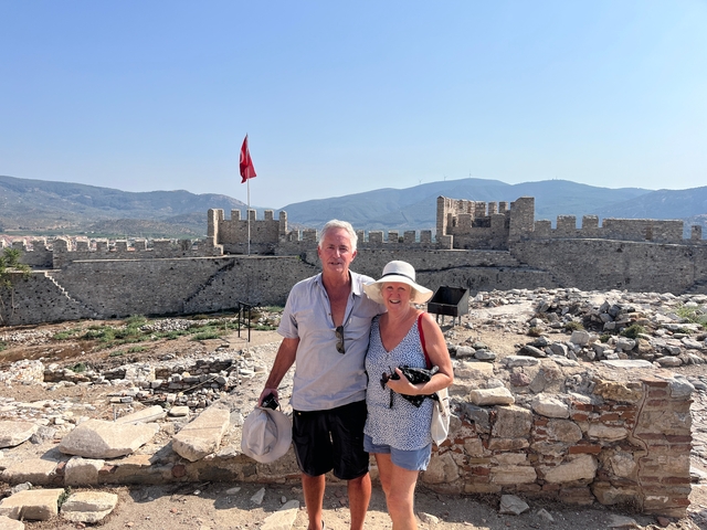 Two people standing in front of a stone fortress with a Turkish flag.