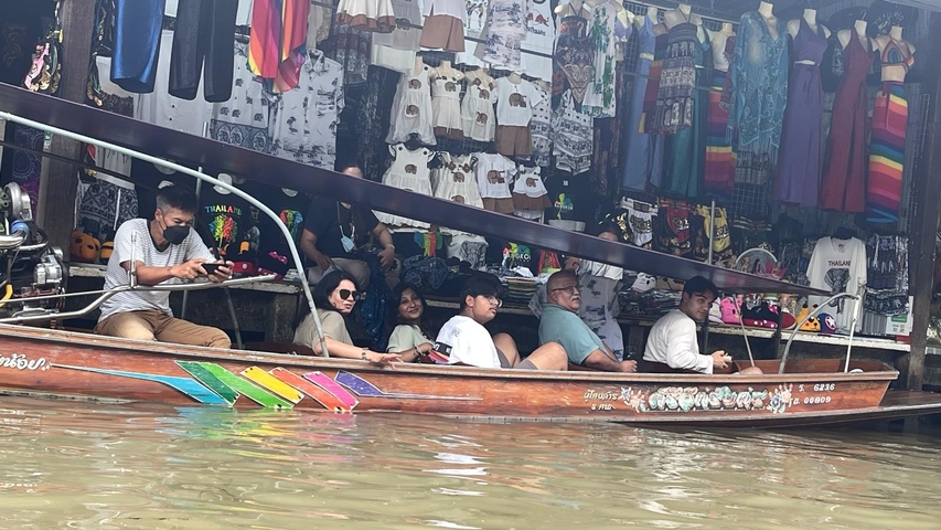       People riding a colorful boat at a floating market with shops in the background.
  