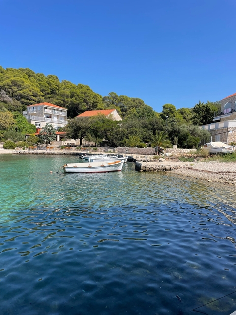 Small boats floating on clear turquoise water near a pebble beach with houses and trees in the background.