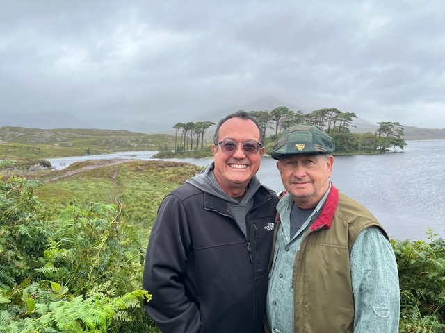       Two people standing in windy conditions with trees and water in the background.
  