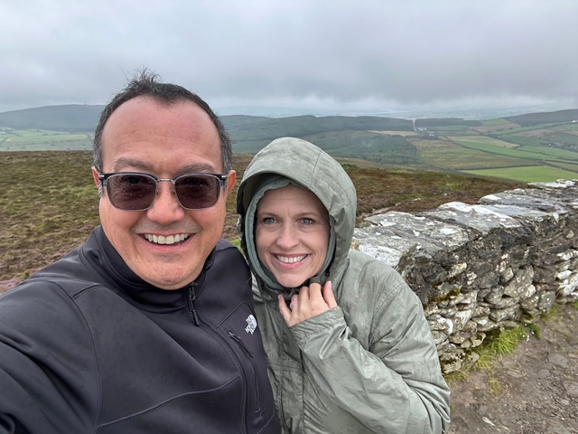       Couple posing on a windy hilltop with expansive countryside views.
  
