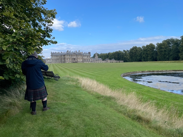       Person in kilt standing near a large mansion and a pond.
  