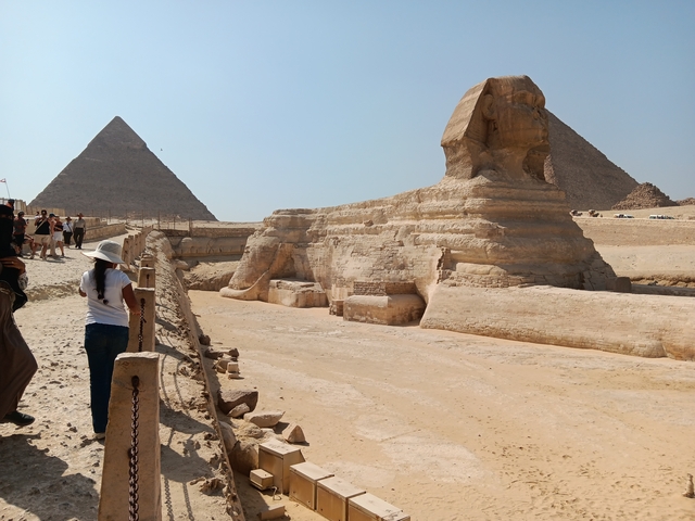 Sphinx in the foreground with two pyramids in the background and tourists walking around.