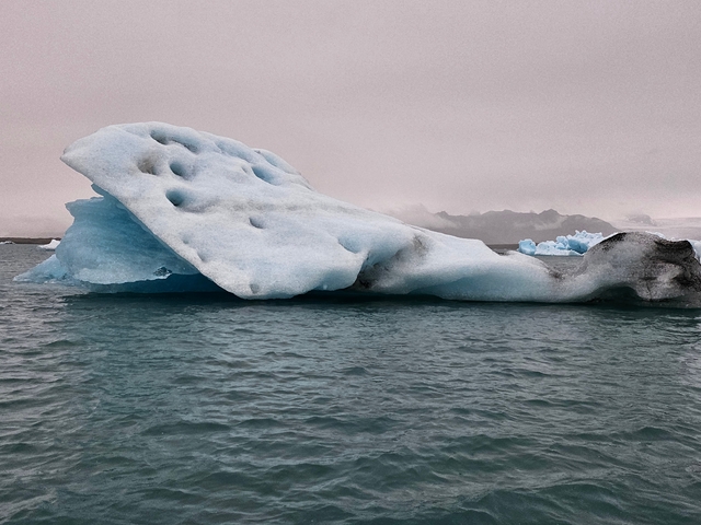 Large blue iceberg floating in the water with cloudy skies.