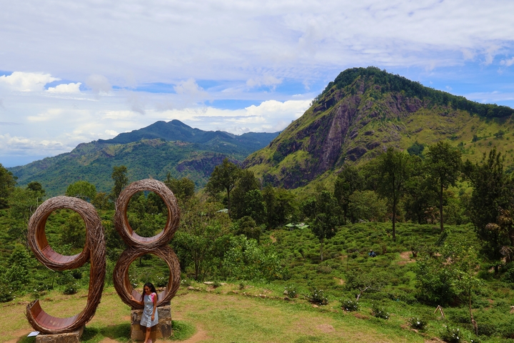 Person posing with a large wooden sculpture of the number 98 with mountains in the background.