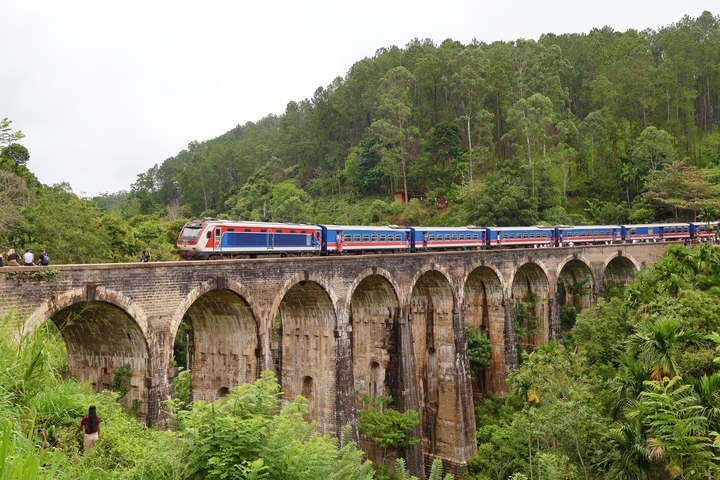 A colorful train crossing a historic nine-arch bridge in a lush landscape.