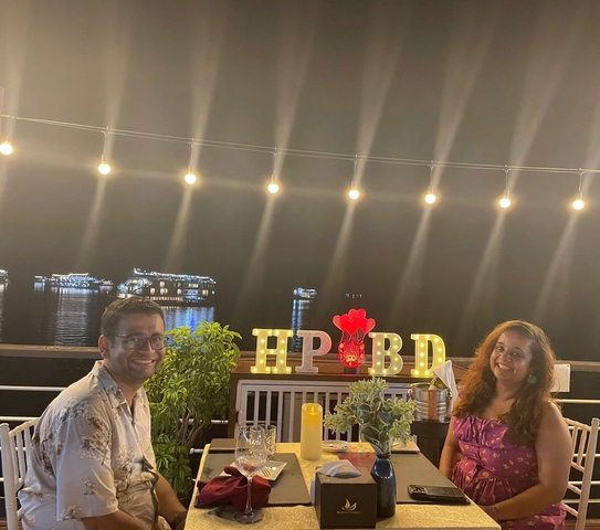 Two people smiling at a decorated table with lights, by a waterfront at night.