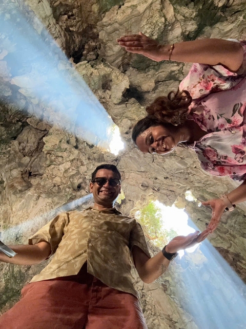 A couple smiling inside a cave with sunlight streaming through openings above.