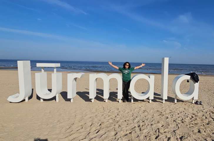       Person posing with large letters spelling 'Jūrmala' on a beach.
  