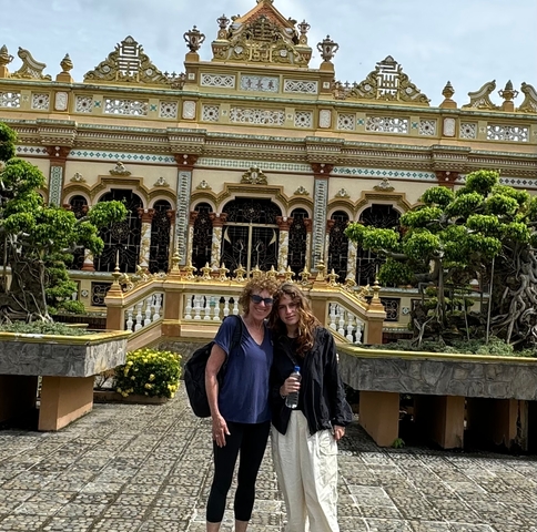 Two people standing in front of a decorative building with ornate architecture and greenery.