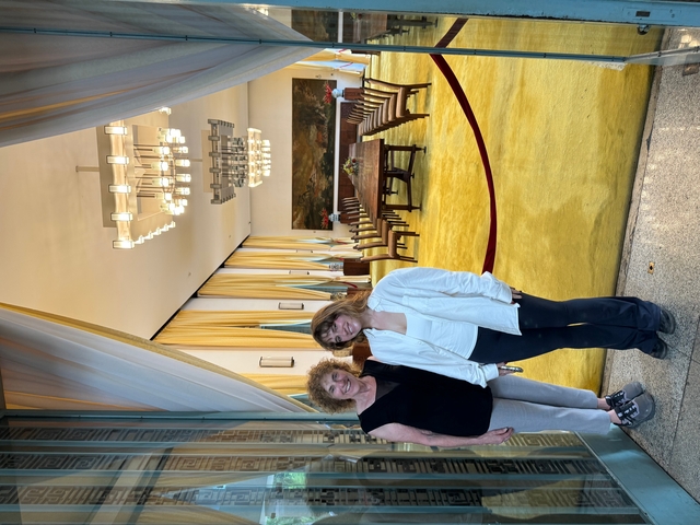 Two women standing in an ornate interior with golden decorations and chandeliers.