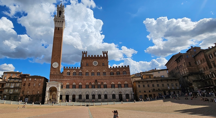 A large square with a historic brick building and a tower under a blue sky.