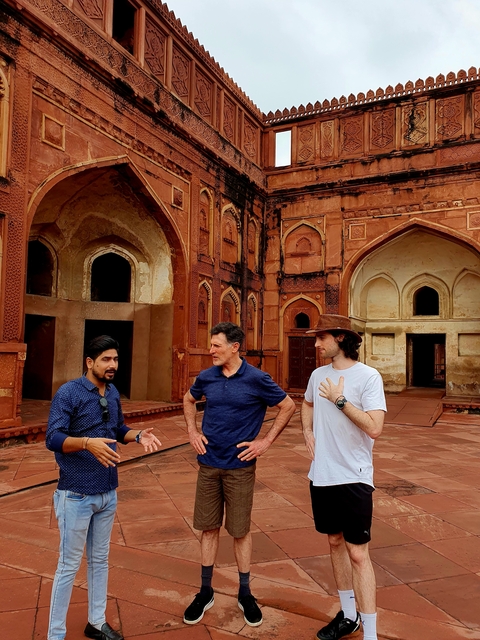       Three men speaking in front of an ancient red building with arches.
  
