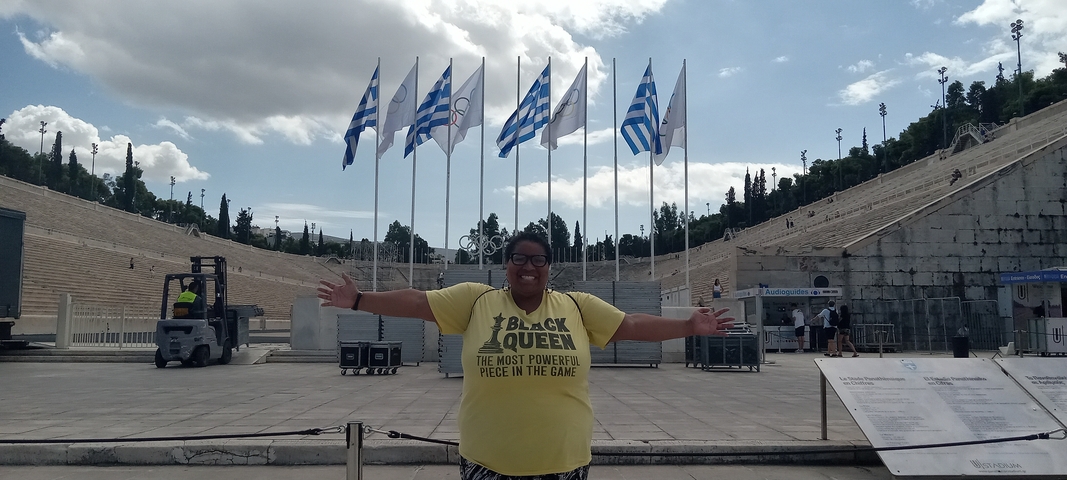 A person in a yellow t-shirt standing with arms open at an ancient stadium.