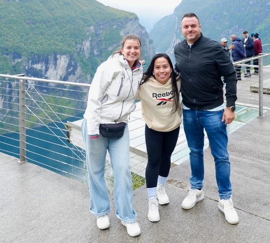 Three people smiling on a viewpoint platform overlooking a fjord.