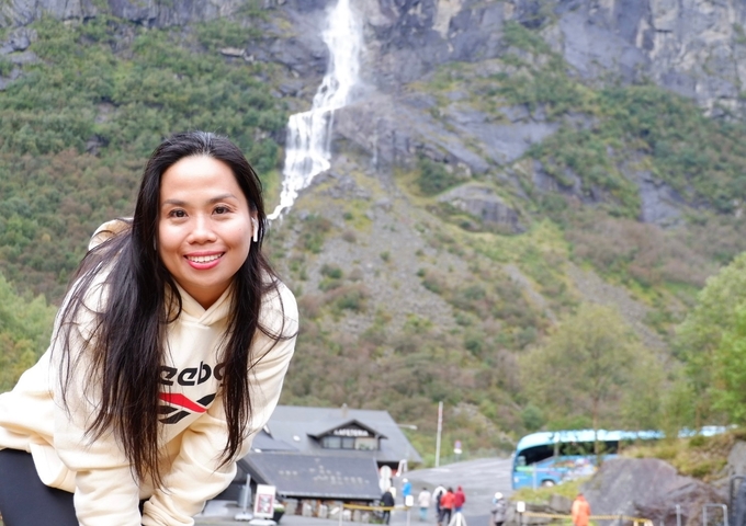 Person posing with a waterfall in the background in a mountainous area.