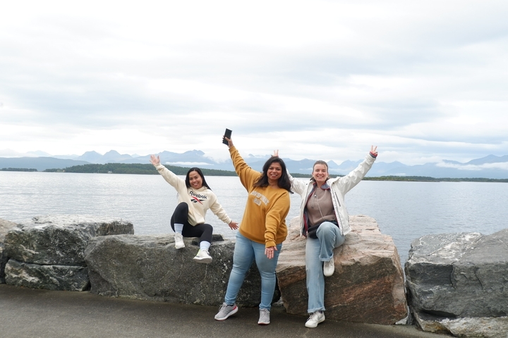 Three people sitting on rocks by a waterfront with mountains in the distance.