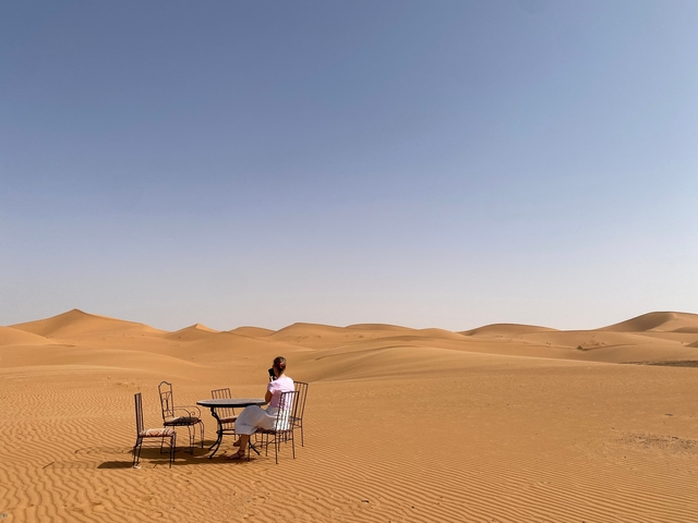 Person sitting at a table set up in the desert with rolling sand dunes.