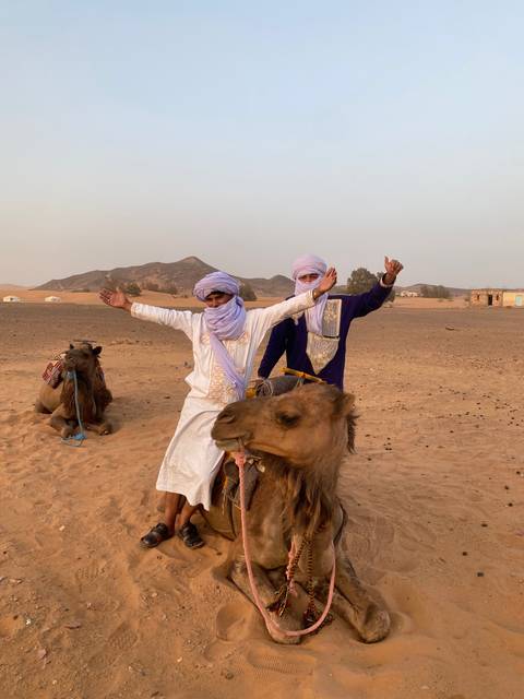 Two people dressed in traditional attire posing with camels in the desert.