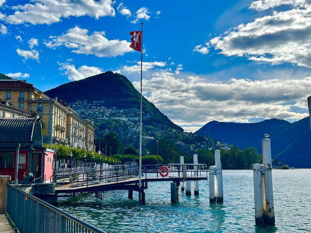 Lugano lake with mountains, a Swiss flag, and pier in the foreground.