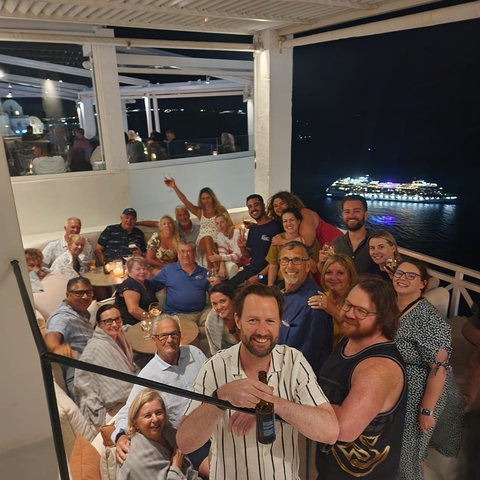 A group of people at a restaurant terrace at night with a cruise ship in the background.